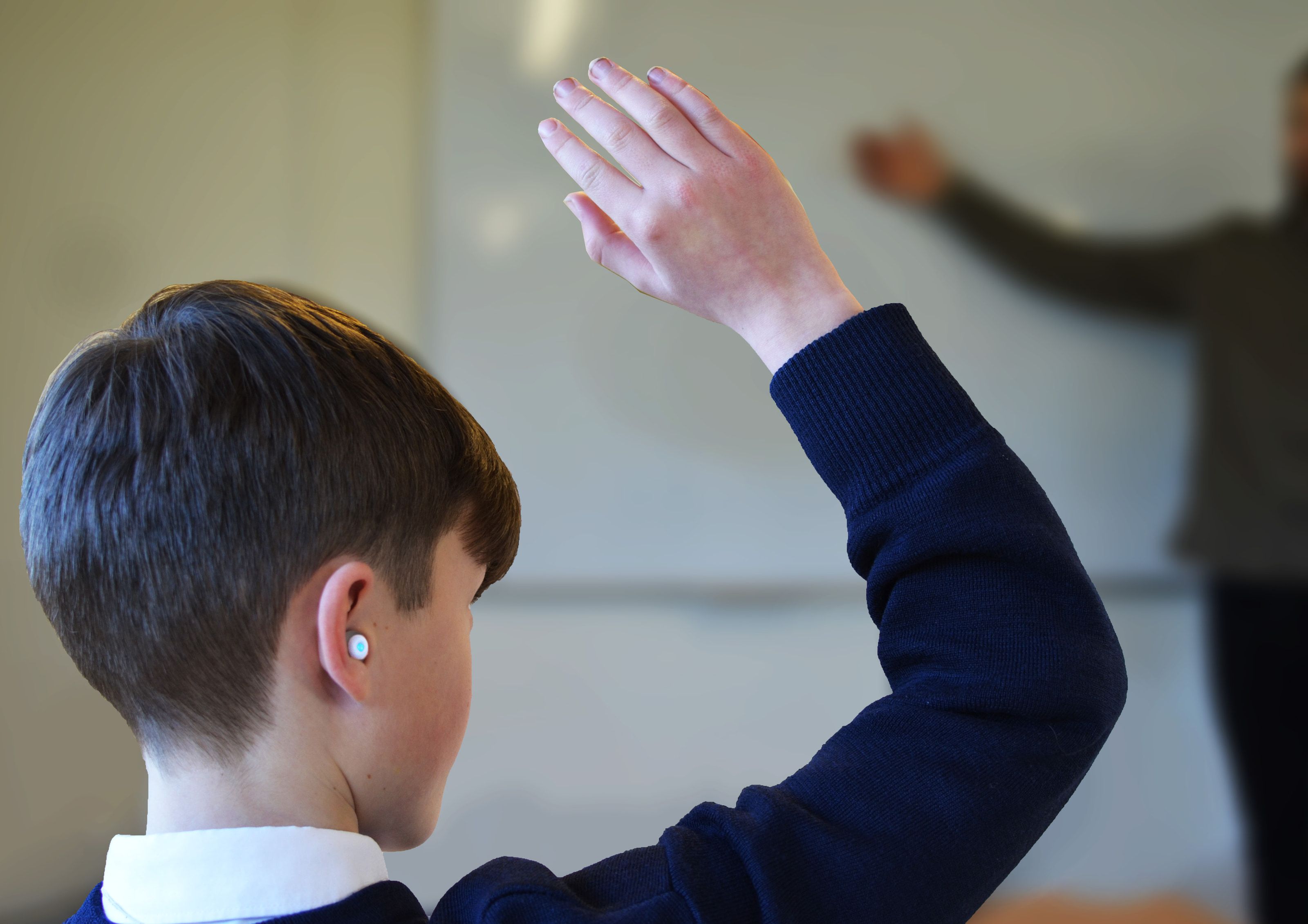 A pupil seen from behind with a hand raised in class, wearing a small white Phonics earbud.