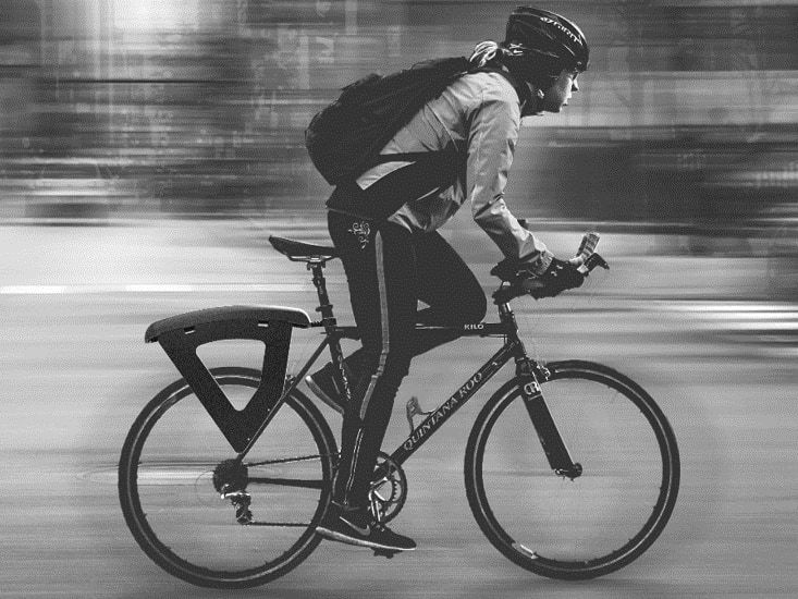 A black-and-white photo of a cyclist riding through a city, with the black pannier rack added to the back of the bike.