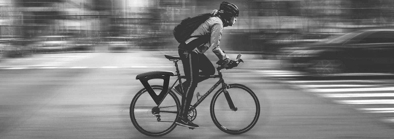 A black-and-white photo of a cyclist riding through a city, with the black pannier rack added to the back of the bike.