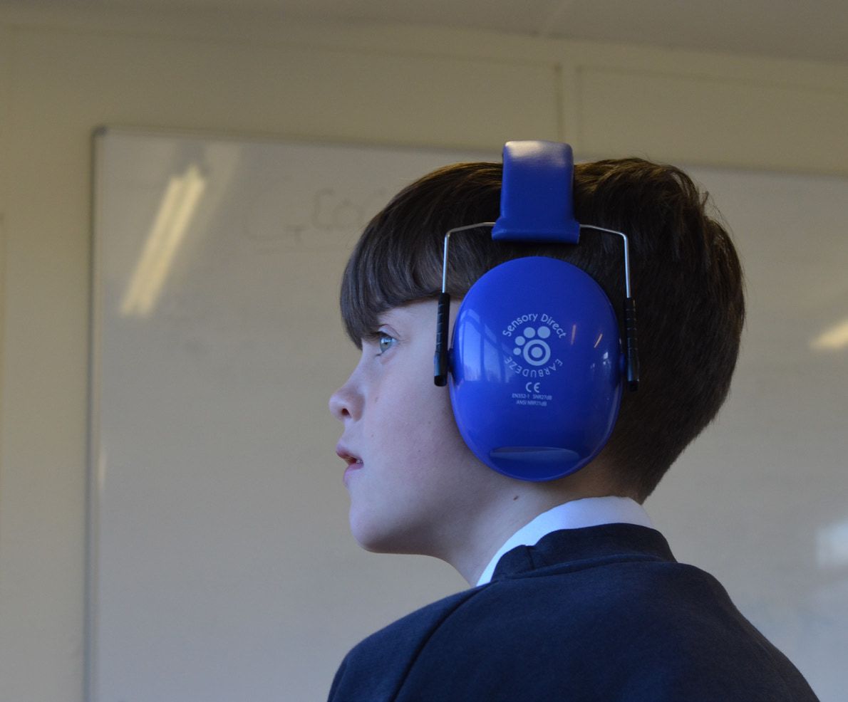 A pupil in profile in a classroom, wearing bulky blue over-ear ear mufflers.