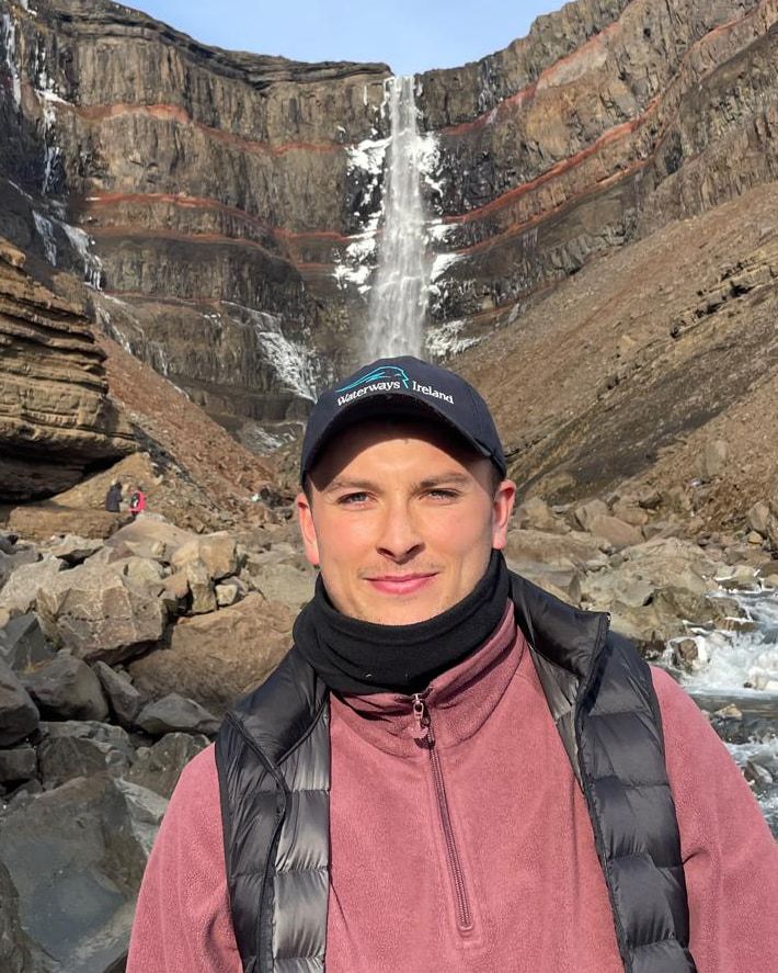 Calum smiling, in a cap and fleece, in front of a tall waterfall falling down layered red and brown cliffs.