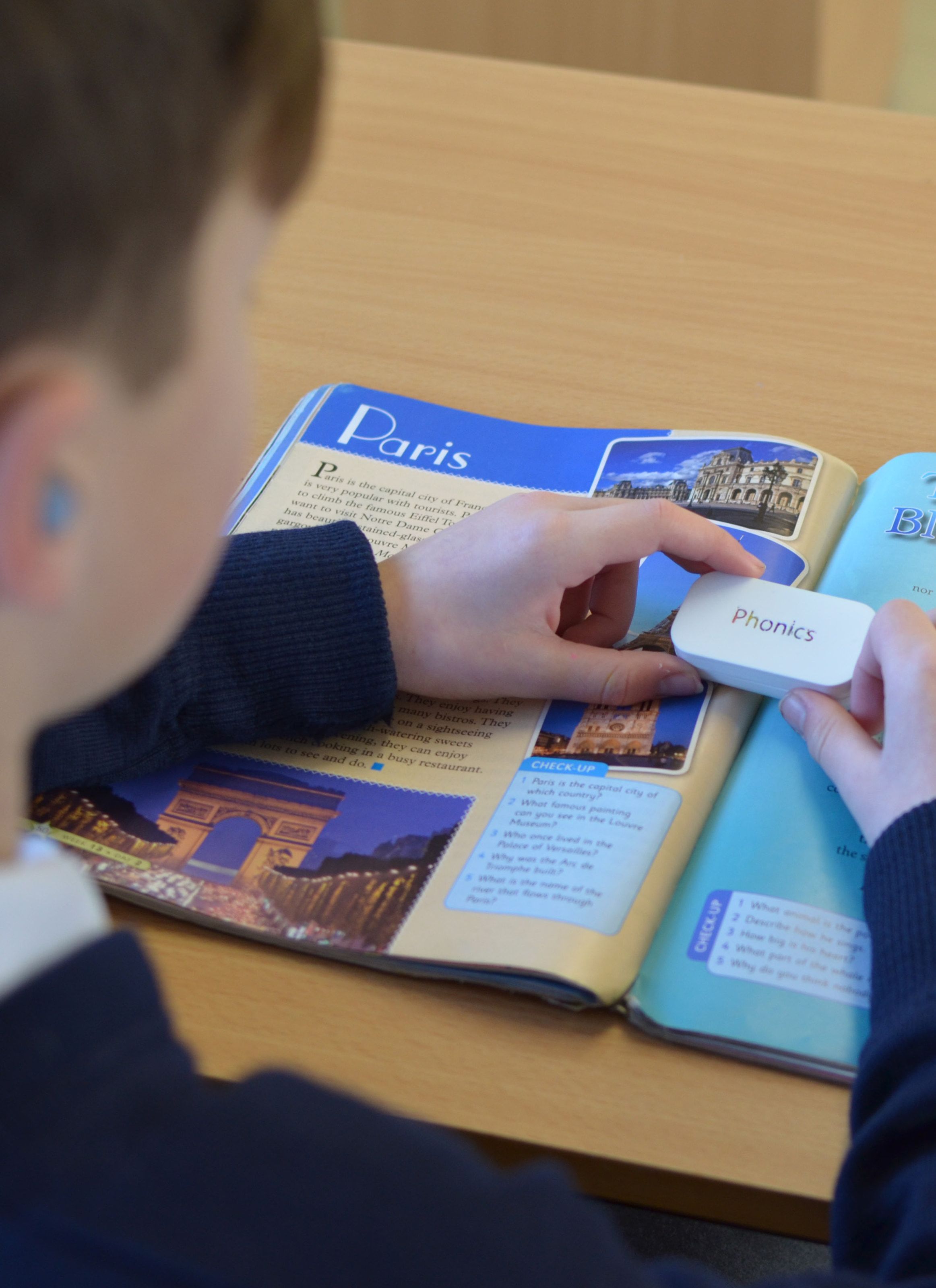 Over the shoulder of a pupil holding the white Phonics case on a textbook page about Paris, with a light blue earbud in their ear.